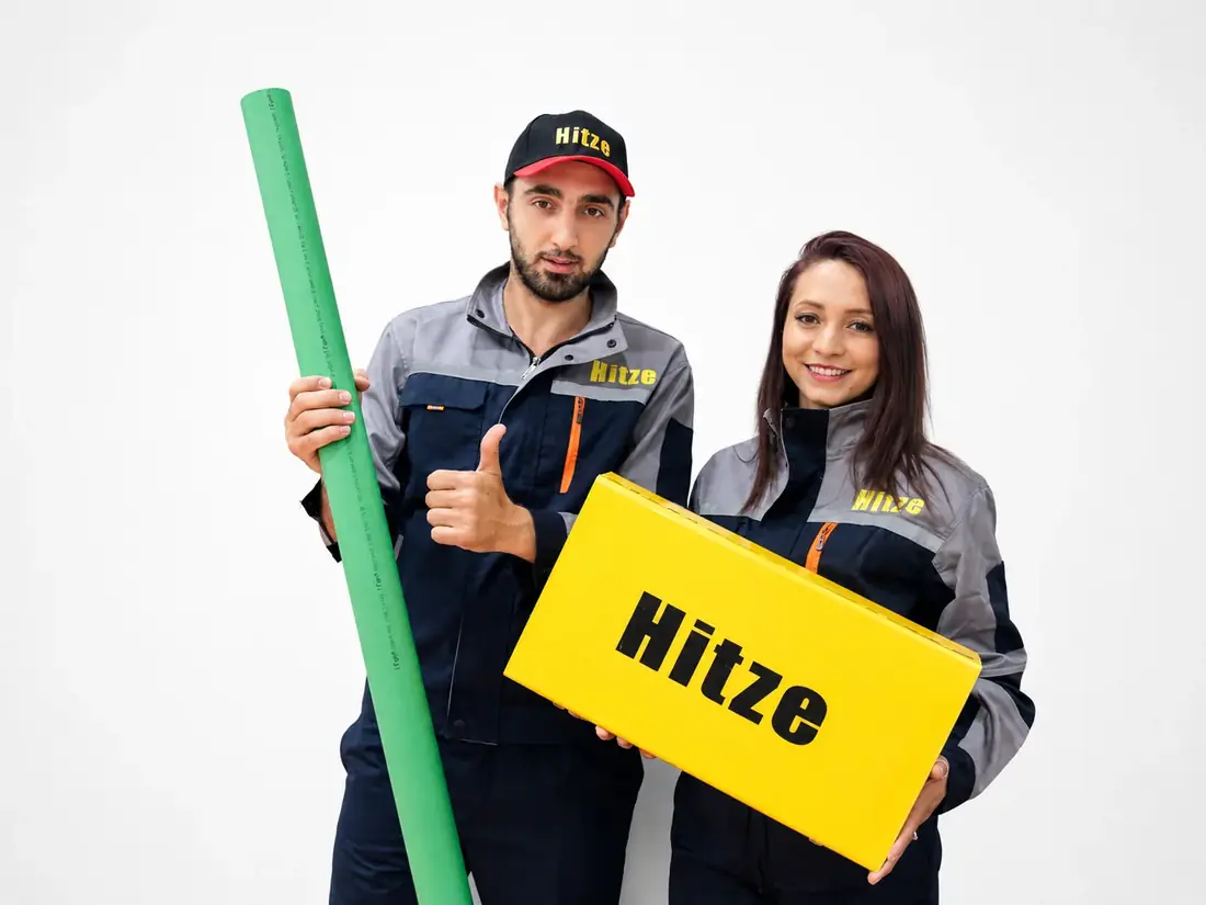 Two Hitze staff in branded grey-and-navy workwear and caps, one holding a green pipe length and giving a thumbs-up, the other holding a yellow Hitze box