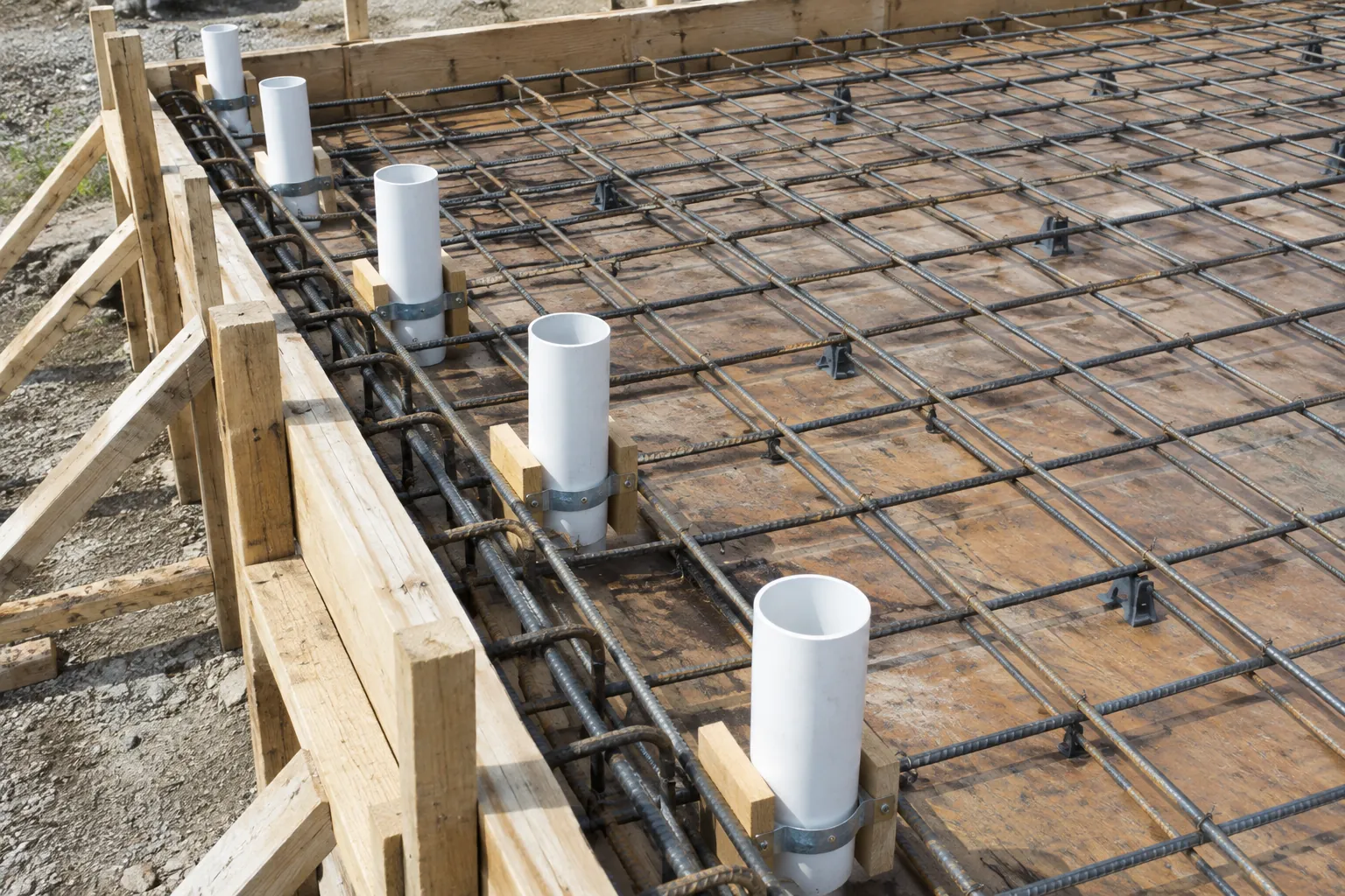 Row of white PVC pipe sleeves clamped upright to timber blocks on slab formwork between steel rebar mesh before the concrete pour